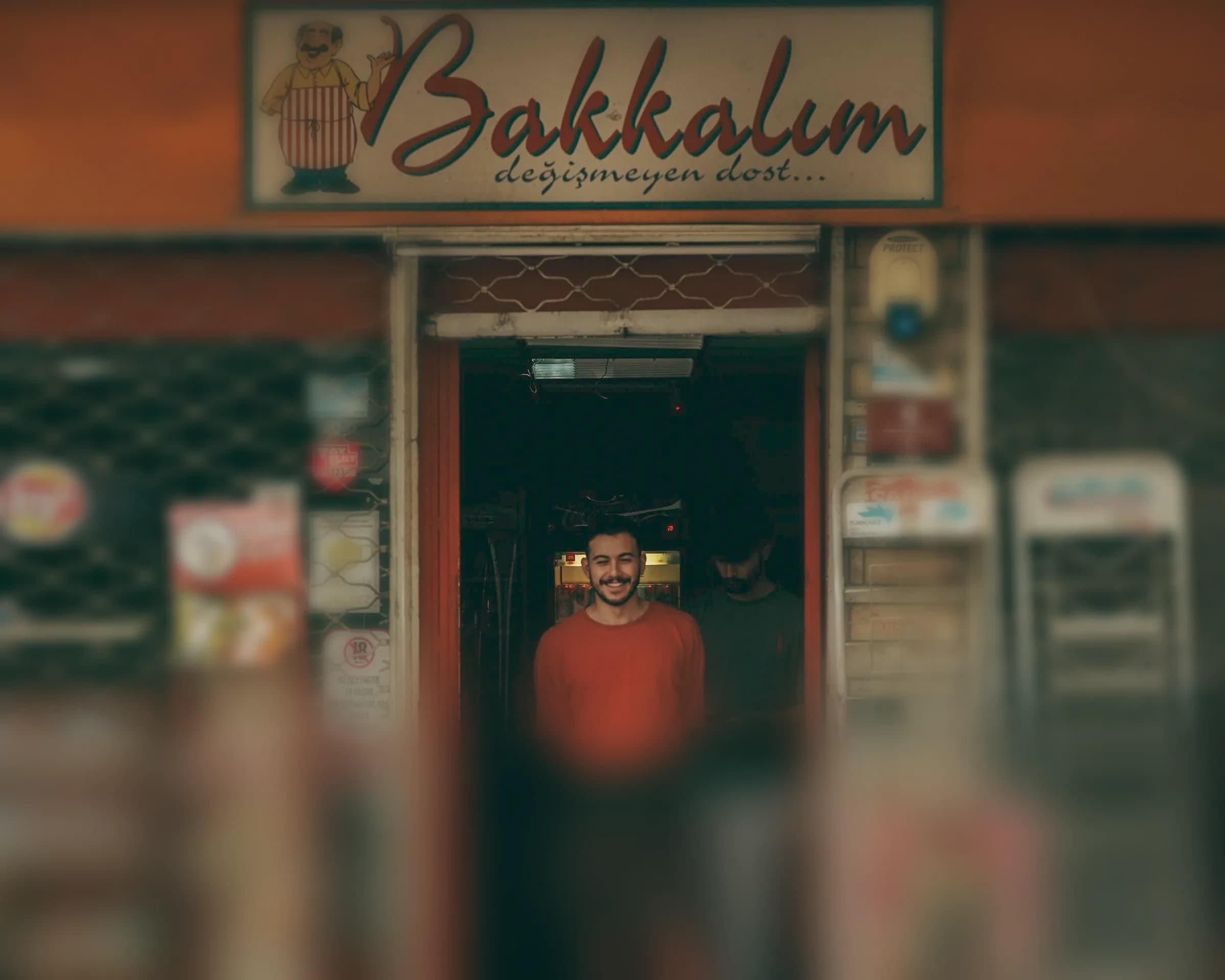 Bakkalım tabelalı mahalle bakkalının kapısında gülümseyen kırmızı tişörtlü genç esnaf