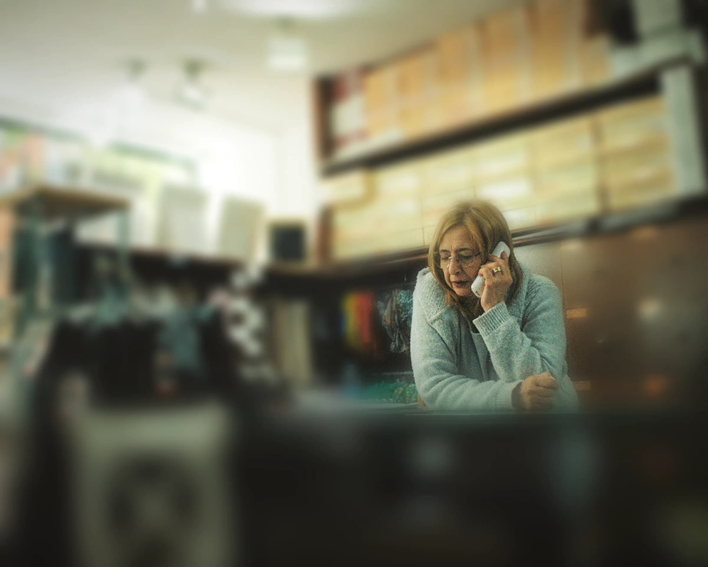 Giyim mağazasında tezgâha yaslanıp telefonla konuşan gözlüklü kadın esnaf, arkada ürün rafları