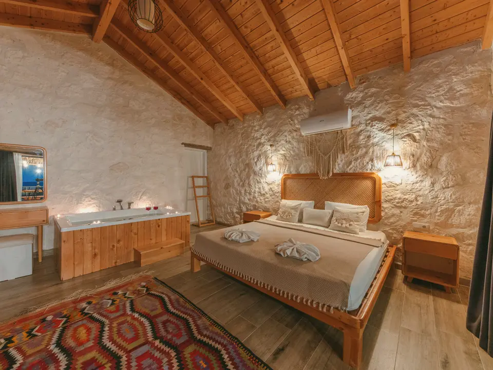 Taş duvarlı, ahşap tavanlı butik otel odasında çift kişilik yatak ve yerde renkli kilim