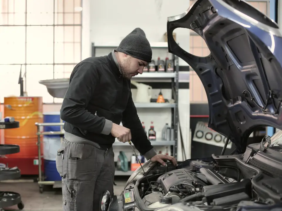 Oto servisinde kaputu açık aracın motor yağını kontrol eden usta, arkada yağ bidonları ve raflar