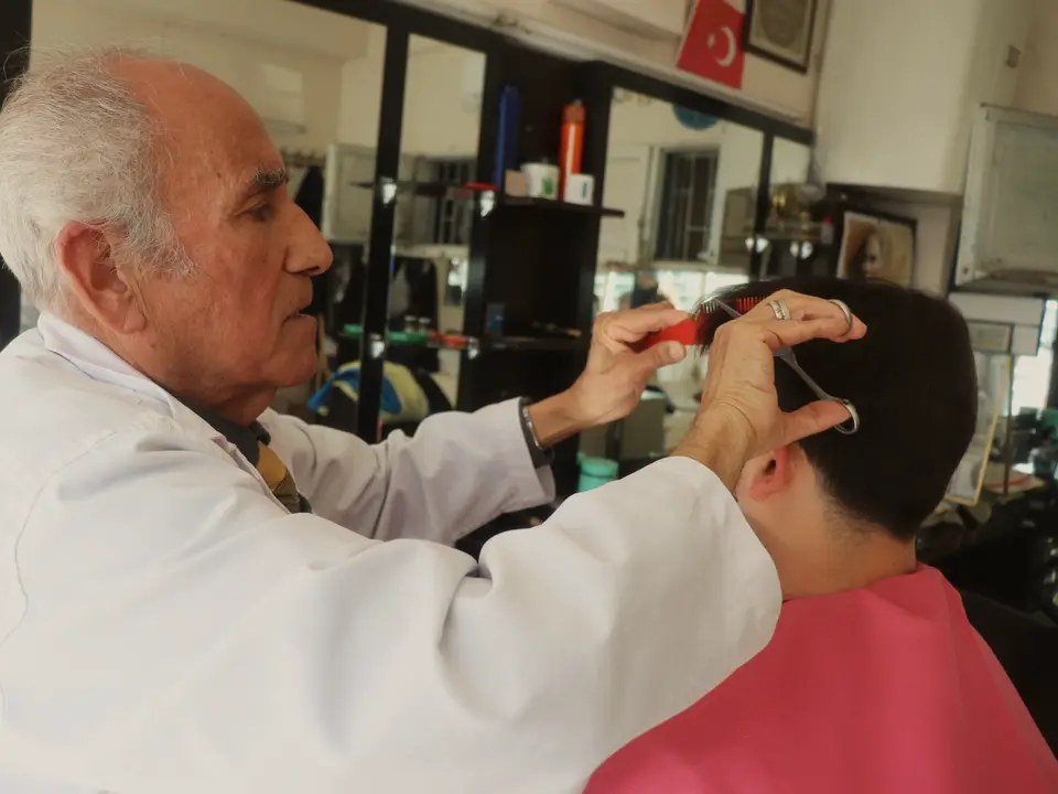 Mahalle berberinde beyaz önlüklü yaşlı berber müşterisinin saçını tarak ve makasla kesiyor