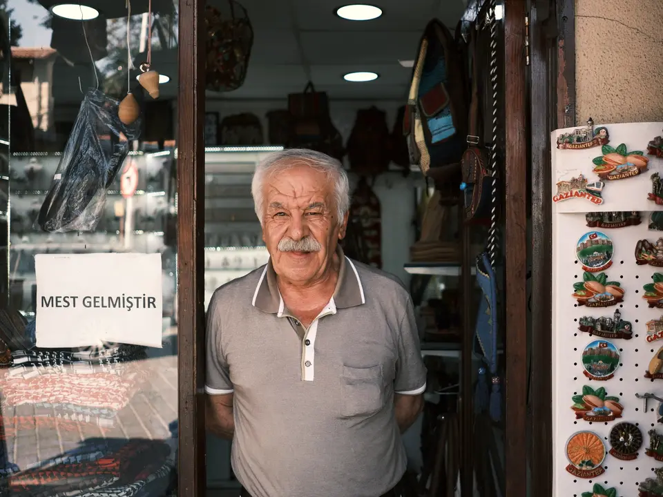Çanta ve hediyelik eşya dükkânının kapısında duran yaşlı esnaf, camda Türkçe yazılmış bir duyuru
